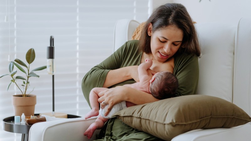 A photograph of a mother in a green shirt breastfeeding her infant. She is sitting in a white armchair next to a small, potted plant in front of a white wall and curtained window.